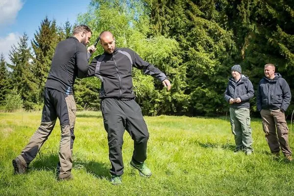 Zwei Personen üben eine Selbstverteidigungstechnik auf einer Wiese, während zwei weitere zuschauen