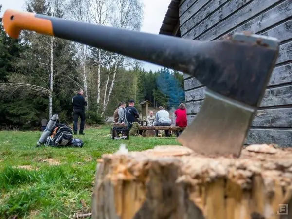 Axt auf Holzscheit, im Hintergrund Gruppe am Tisch vor Holzhaus