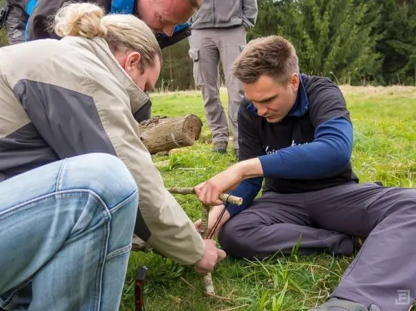 Drei Personen arbeiten gemeinsam an einer Holzarbeit mit einem Stock und einem weiteren Werkzeug