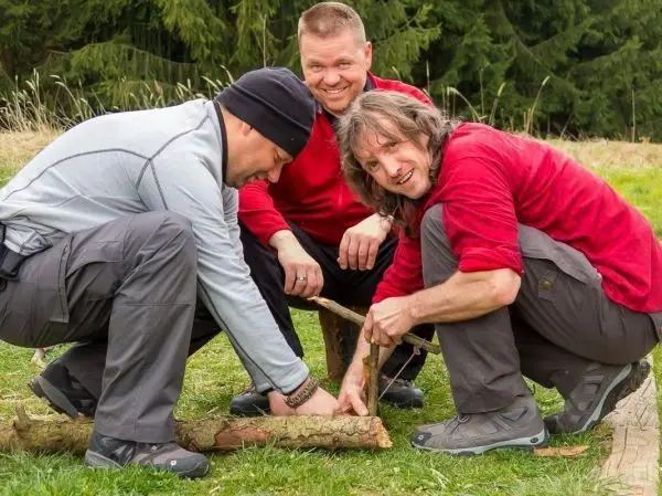 Drei Personen bearbeiten einen Holzstock auf dem Boden beim Überlebenstraining