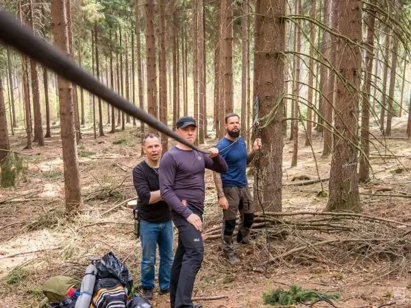 Drei Personen halten eine lange Stange in einem Wald zwischen Bäumen