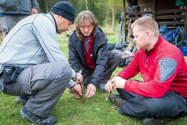 Drei Personen üben das Zundern mit Holzstücken auf dem Boden