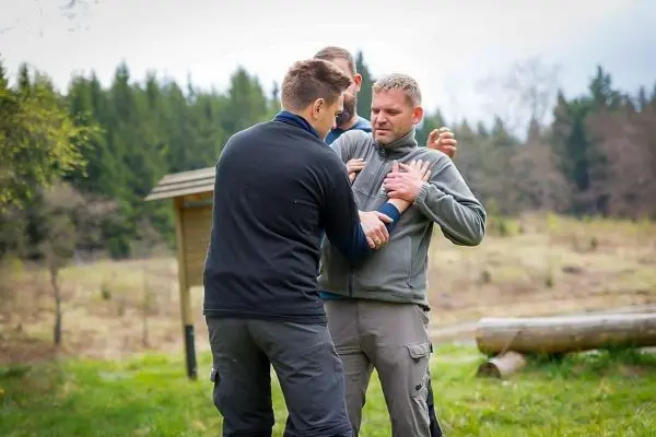 Drei Personen üben eine körperliche Technik zur Selbstverteidigung im Freien