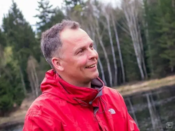 Ein Mann in roter Jacke lächelt am Ufer eines Gewässers