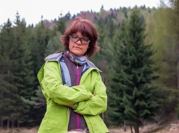 Eine Person in einer grünen Jacke steht mit verschränkten Armen vor einem Wald