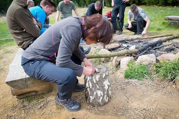 Frau bearbeitet Holzstück mit einem Messer neben einem Lagerfeuer