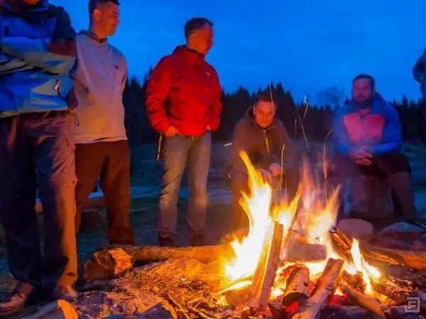 Gruppe von Personen sitzt um ein Feuer und beobachtet die Flammen