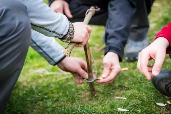 Hände binden eine Schnur um einen Stock auf dem Gras beim Überlebenstraining