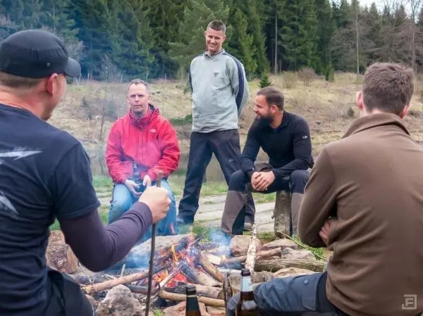 Kreis von Personen sitzt um ein Lagerfeuer mit Holzscheiten und rauchenden Flammen