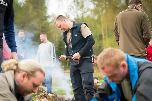 Mann bearbeitet Holzstück mit einem Messer, Rauch steigt von einem kleinen Feuer auf