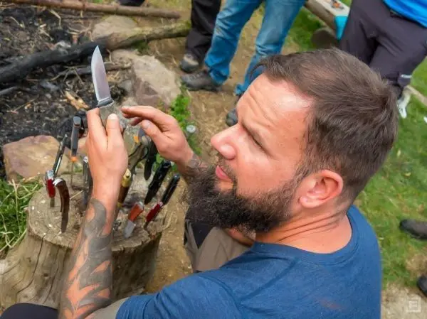 Mann hält ein Messer und betrachtet die Klinge, umgeben von weiteren Messern auf einem Baumstumpf