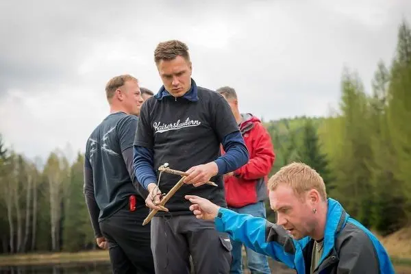 Mann hält selbstgebaute Feuerbohrer-Konstruktion, während andere zuschauen