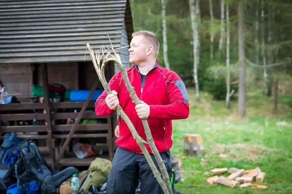 Mann hält zwei Äste in der Hand, im Hintergrund eine Holzstruktur