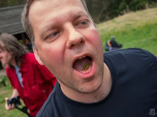 Mann mit einer kleinen Frucht im Mund, im Hintergrund eine Person mit Kamera