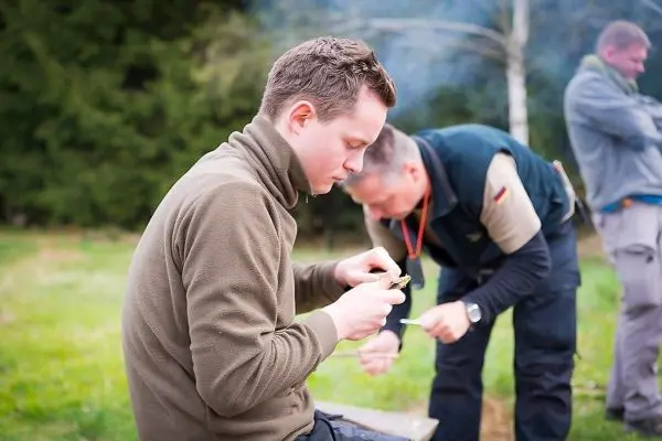Mann schnitzt mit einem Messer an einem Holzstück, während ein anderer im Hintergrund arbeitet