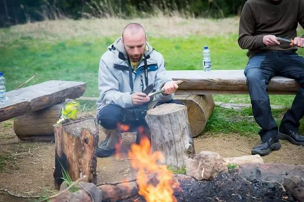 Mann schnitzt mit einem Messer Holzstück neben einem Lagerfeuer