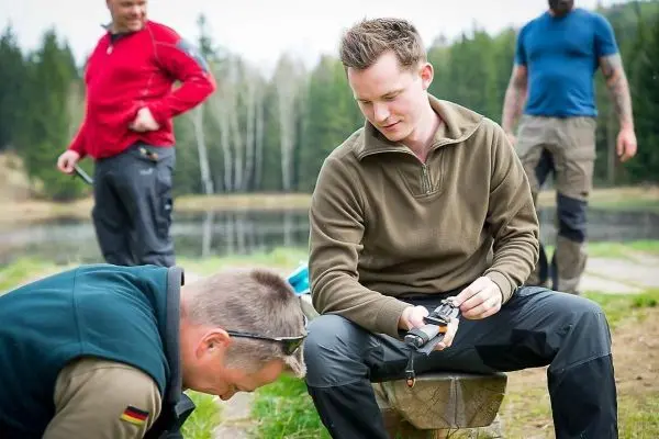 Mann sitzt auf Holzscheit und hält ein Messer, während ein anderer Mann sich bückt