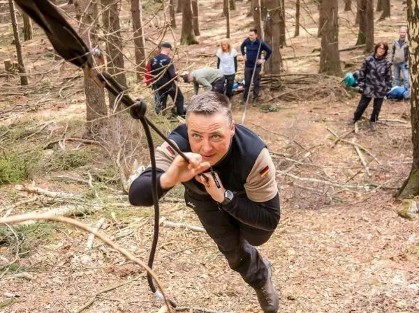 Mann überquert selbstgebaute Seilbrücke im Wald, im Hintergrund weitere Personen