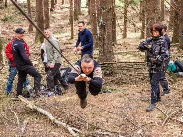 Mann überquert selbstgebaute Seilbrücke im Wald, umgeben von weiteren Personen
