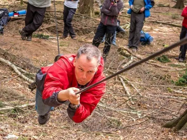 Mann überquert selbstgebaute Seilbrücke im Wald, zieht sich an einem Seil hoch