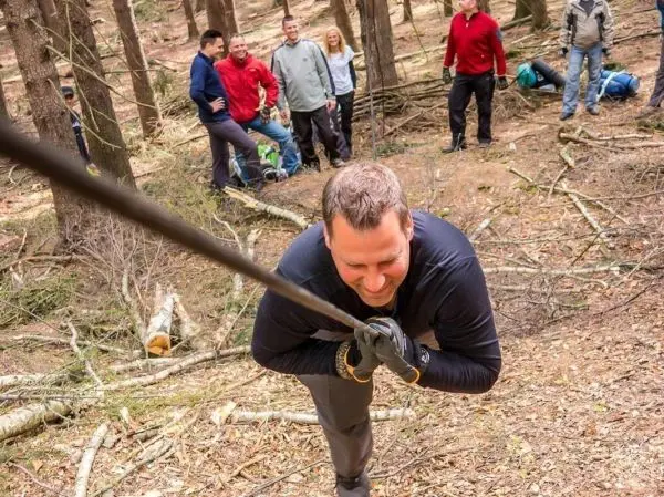 Mann zieht an einem Seil in einem Wald, Zuschauer stehen im Hintergrund