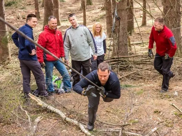 Mann zieht an einem Seil, während andere zuschauen und im Hintergrund Äste liegen