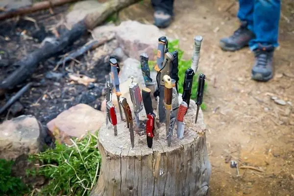 Mehrere Messer stecken in einem Baumstumpf neben einem kleinen Lagerfeuer