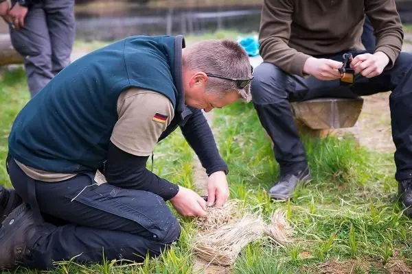 Person kniet und bearbeitet Pflanzenmaterial auf dem Boden beim Überlebenstraining