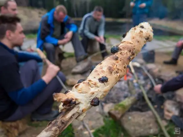 Stock mit gebackenem Brot über Feuer gehalten, im Hintergrund sitzen Personen am Lagerfeuer