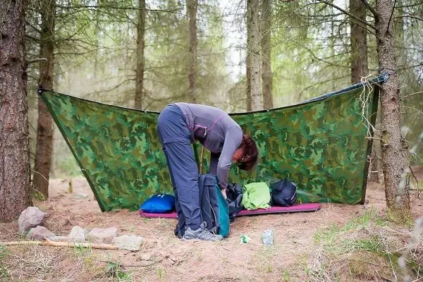 Tarp-Notunterkunft aus Tarnmaterial zwischen Bäumen, Person packt Rucksack