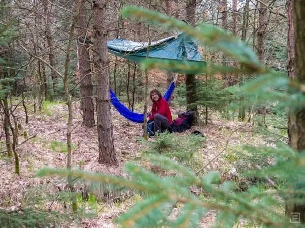 Tarp-Notunterkunft hängt zwischen Bäumen, darunter sitzt eine Person in einer Hängematte