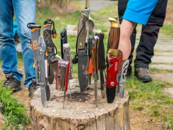 Verschiedene Messer stecken in einem Baumstumpf, während eine Hand ein weiteres Messer hält