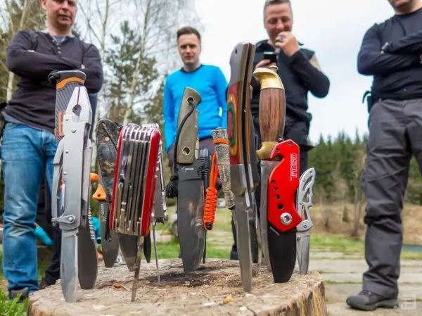Verschiedene Messer und Werkzeuge stecken in einem Baumstumpf, während Personen im Hintergrund beobachten