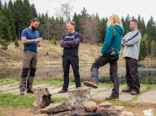 Vier Personen stehen am Ufer eines Gewässers, Holzscheite liegen am Boden