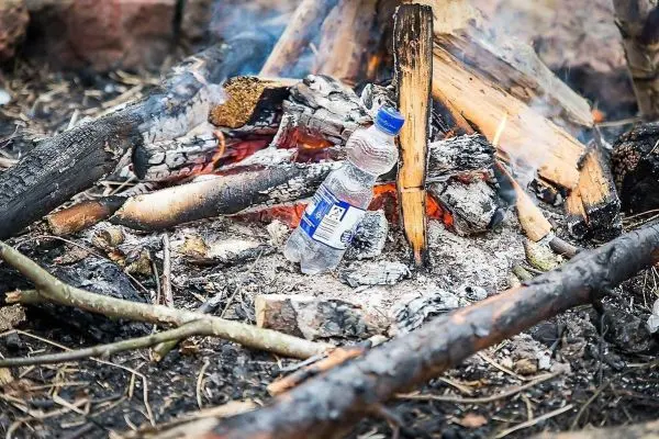 Wasserflasche steht neben glühenden Holzresten eines kleinen Bodenfeuers