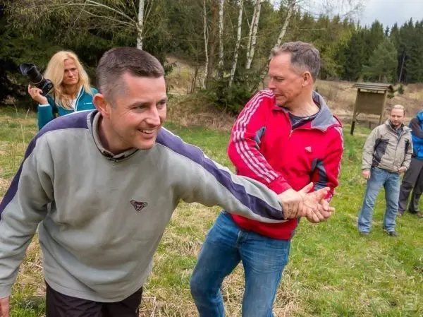 Zwei Männer ziehen an den Händen des anderen in einer Wiese, im Hintergrund stehen weitere Personen