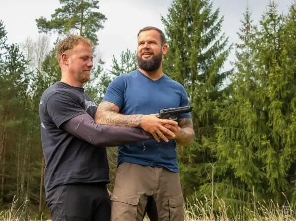 Zwei Personen stehen im Wald, eine hält eine Pistole, die andere greift unterstützend zu