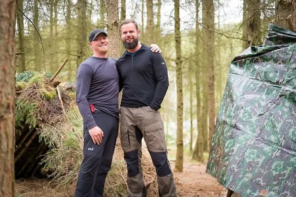 Zwei Personen stehen lächelnd vor einer selbstgebauten Unterkunft im Wald