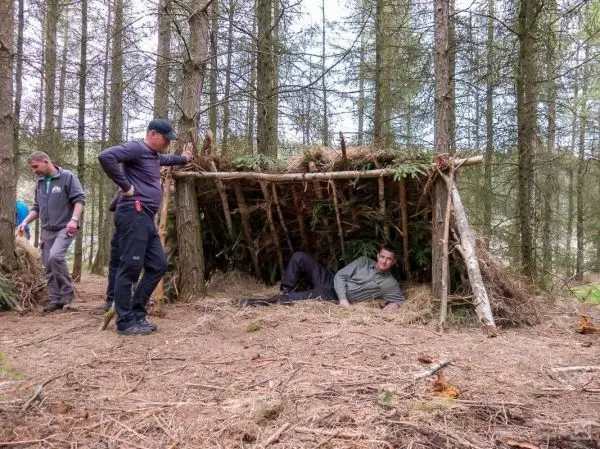 Zwei Personen stehen neben einer selbstgebauten Tarp-Notunterkunft aus Ästen und Zweigen