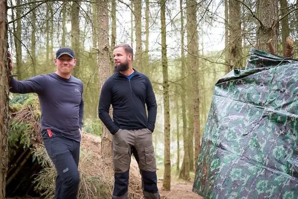 Zwei Personen stehen neben einer Tarp-Notunterkunft im Wald
