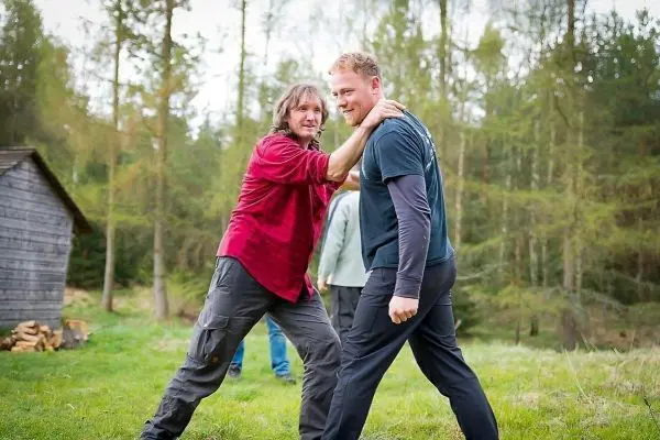 Zwei Personen üben eine körperliche Technik im Freien, umgeben von Bäumen und einer Holzhütte