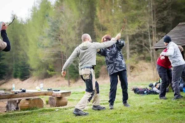 Zwei Personen üben mit Stöcken in einer Wiese, im Hintergrund Ausrüstung sichtbar