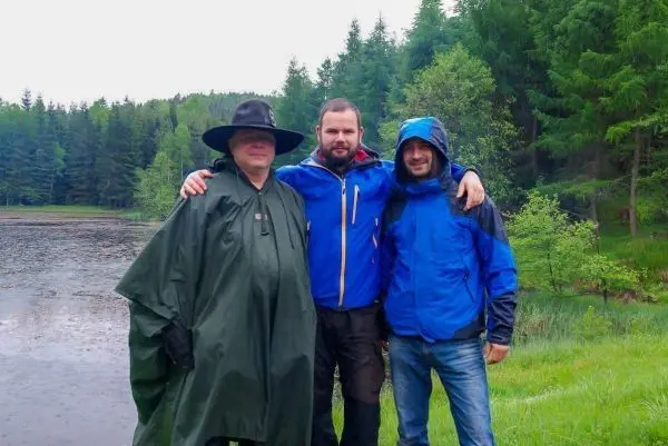 Drei Personen stehen am Ufer eines Gewässers, umgeben von Bäumen und Gras