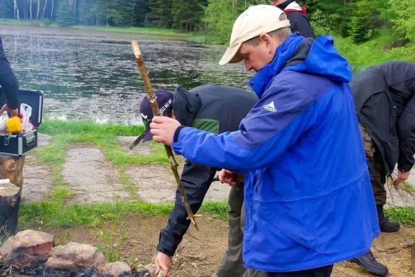 Mann hält Stock über kleines Bodenfeuer am Ufer eines Gewässers