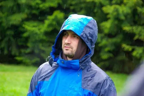 Mann in blauer Regenjacke steht im Regen vor grünen Bäumen