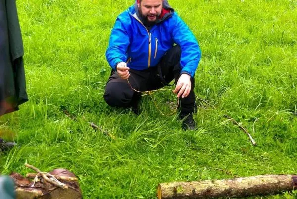 Mann kniet im Gras und bearbeitet einen Stock mit einem Messer