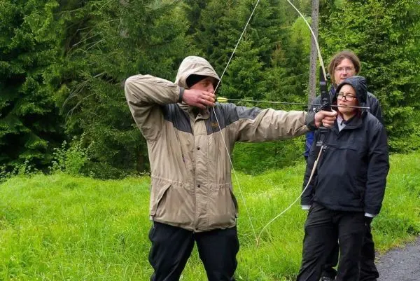 Person spannt einen Bogen und zielt auf ein Ziel, zwei Personen beobachten