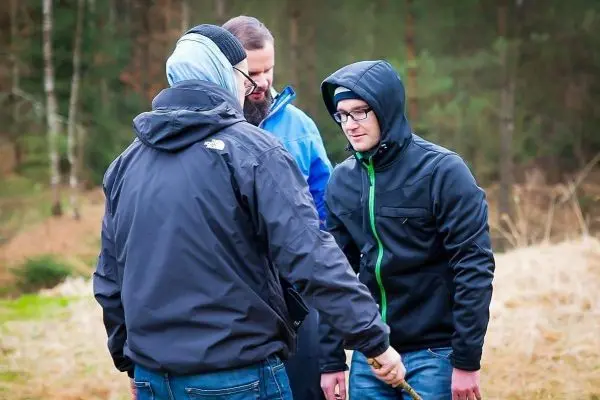 Drei Personen stehen in einer Waldlandschaft und betrachten einen Stock