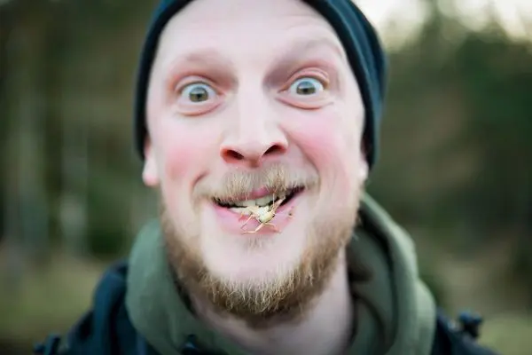 Ein Mann mit Bart lächelt breit und hat ein Insekt im Mund