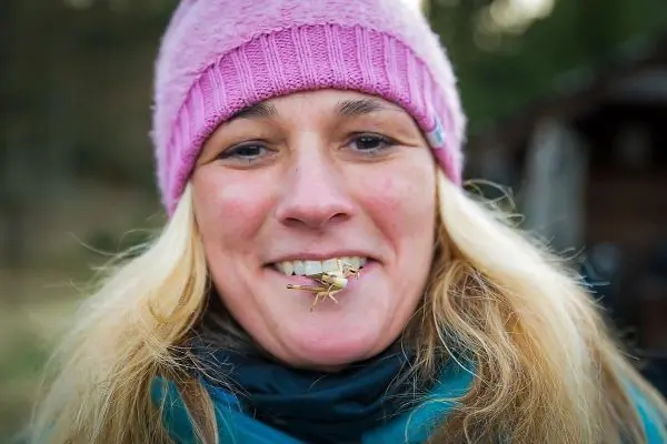 Frau mit blonder Haarpracht und rosa Mütze hält eine Heuschrecke im Mund